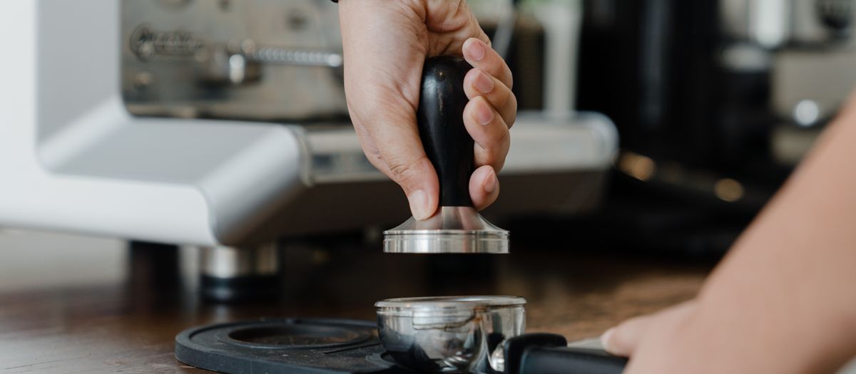 Unrecognizable female bartender pressuring ground coffee in portafilter with tamper for preparing tasty aromatic espresso during work in coffee house